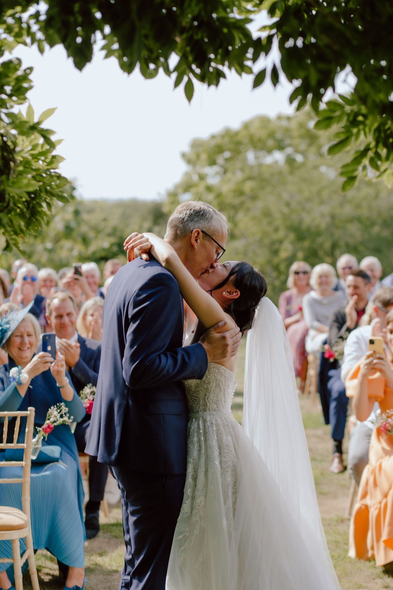 An outdoor wedding ceremony with guests watching a couple share their first kiss under leafy trees.