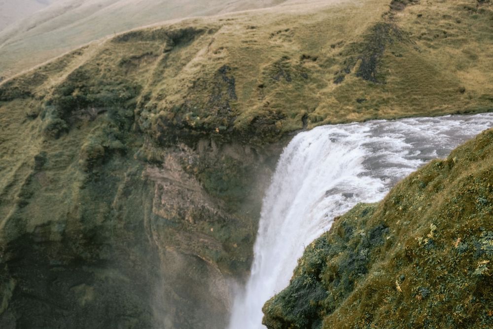 Powerful waterfall cascades down moss-covered cliffs in a mountainous landscape.