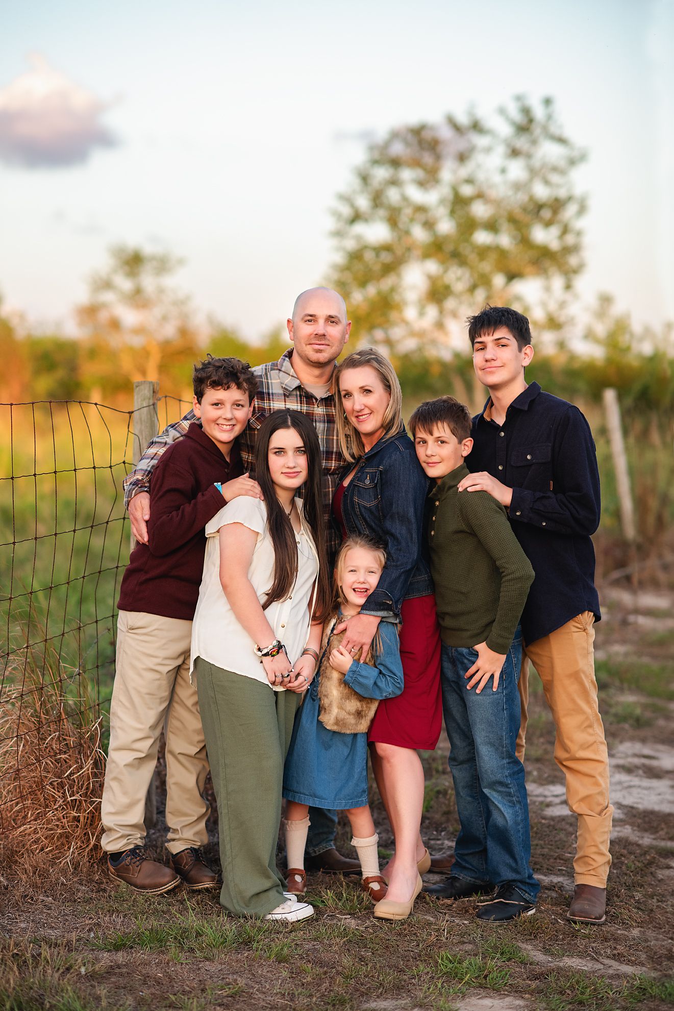 Large family of seven posing together near a fence in a golden sunset outdoor setting.