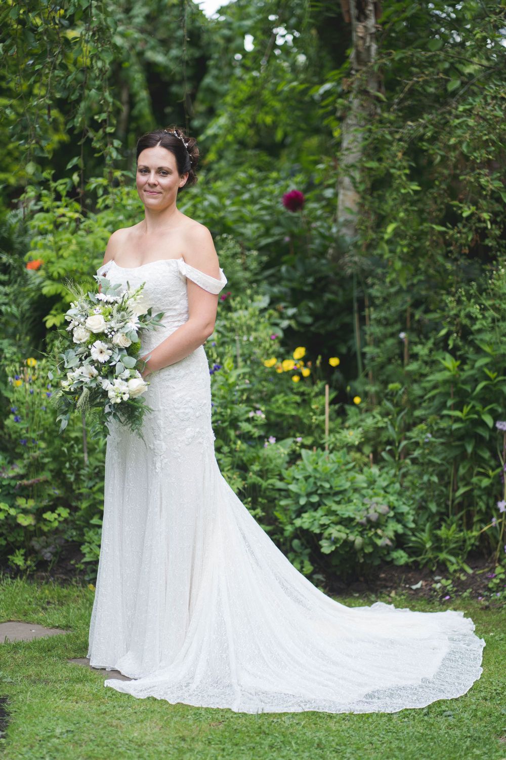 A bride in an elegant off-shoulder wedding gown poses in a vibrant garden setting.