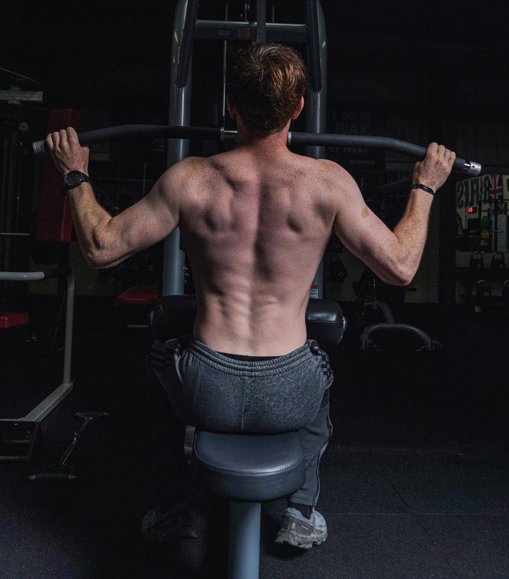 Back view of muscular person performing lat pulldown exercise in dark gym setting.