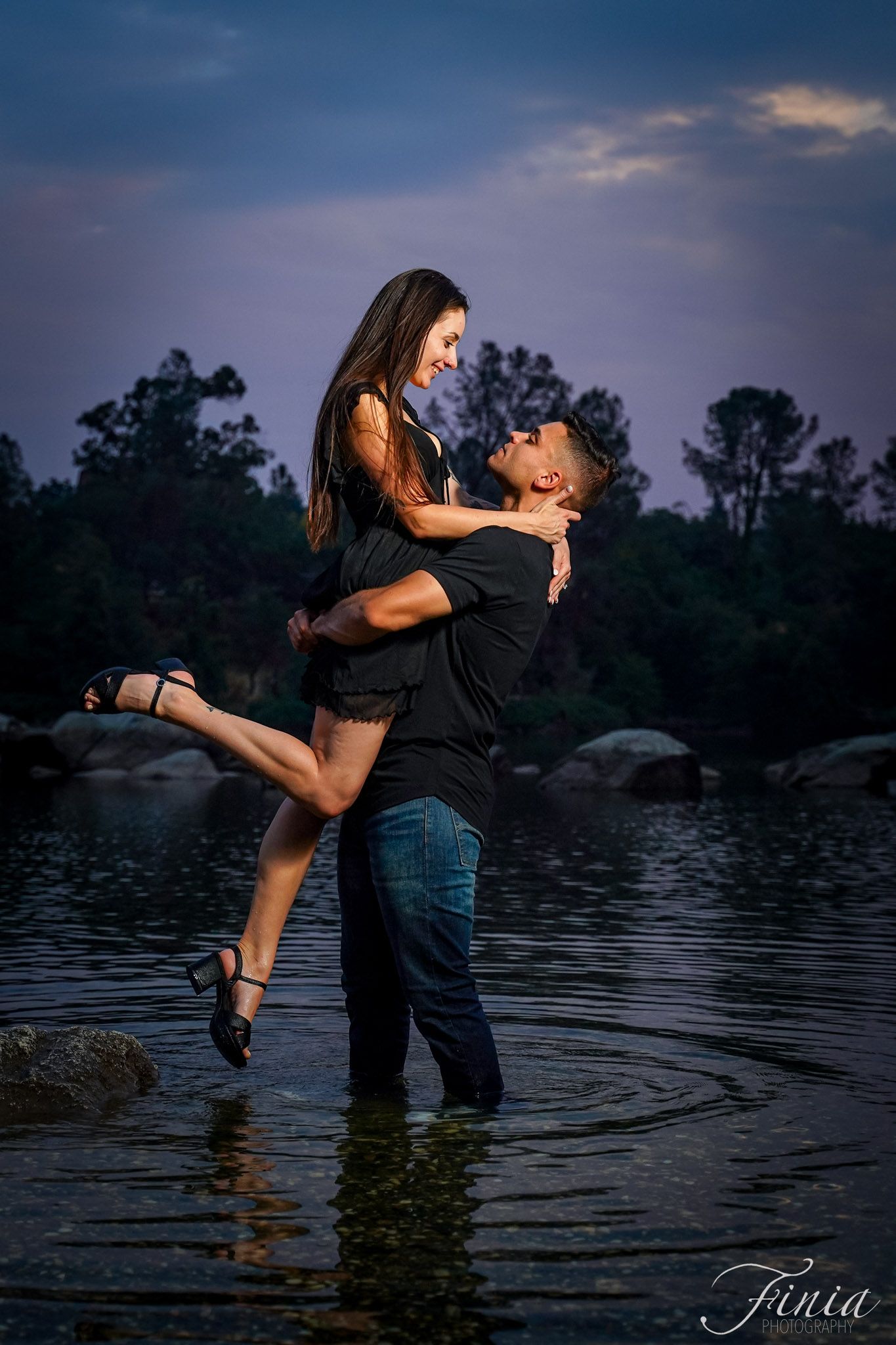 A romantic couple embraces while standing in shallow water at dusk with a dramatic purple sky and trees in background.