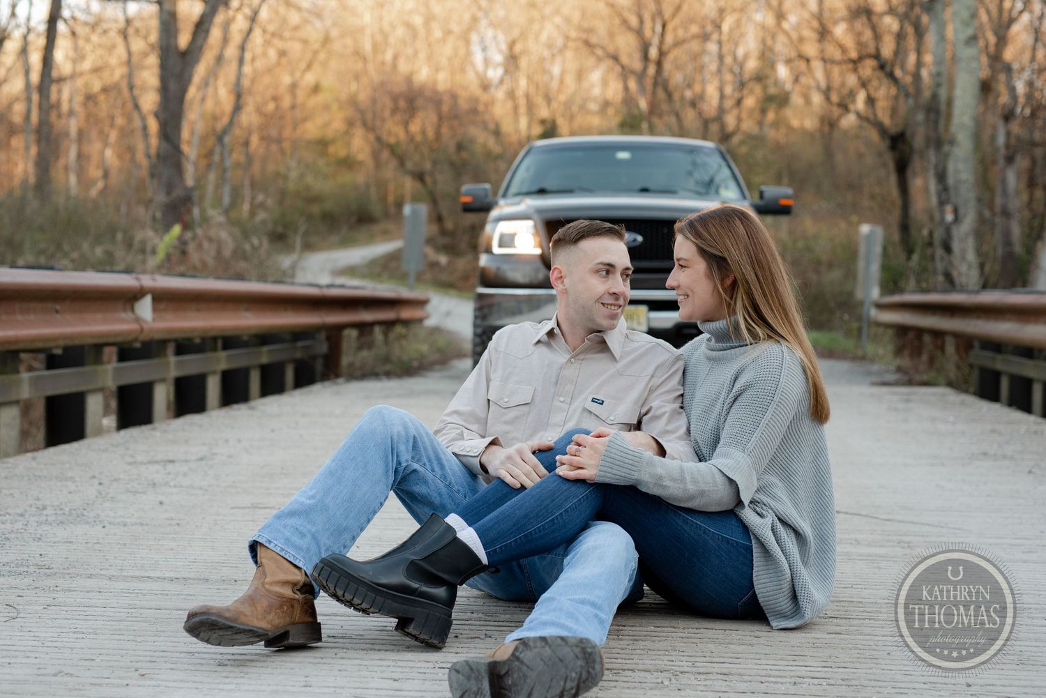 Relaxed couple sitting on wooden bridge during sunset photoshoot with truck in background.