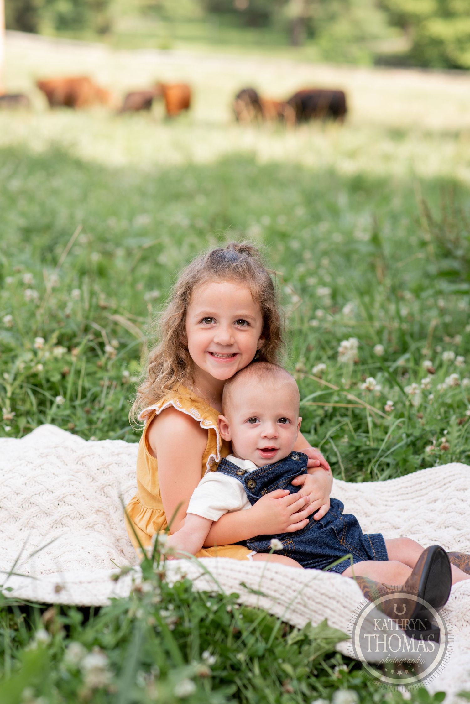 Two children sitting on a white blanket in a field with horses grazing in the background.