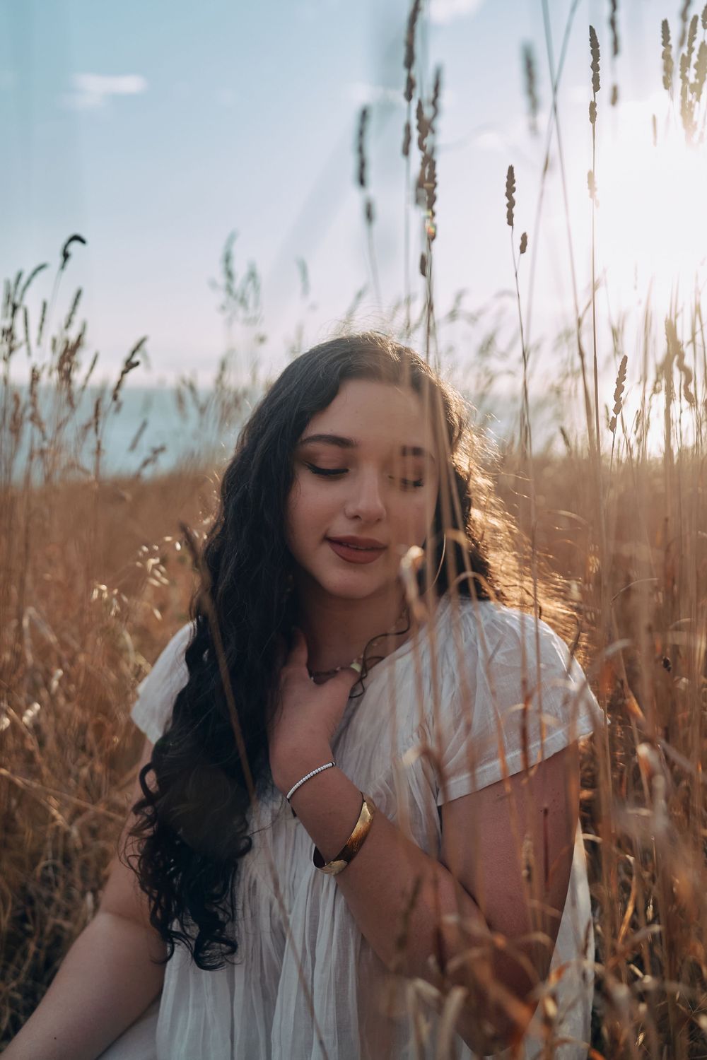 Young woman with dark curly hair in white dress standing in golden wheat field with sunlight glowing behind her.