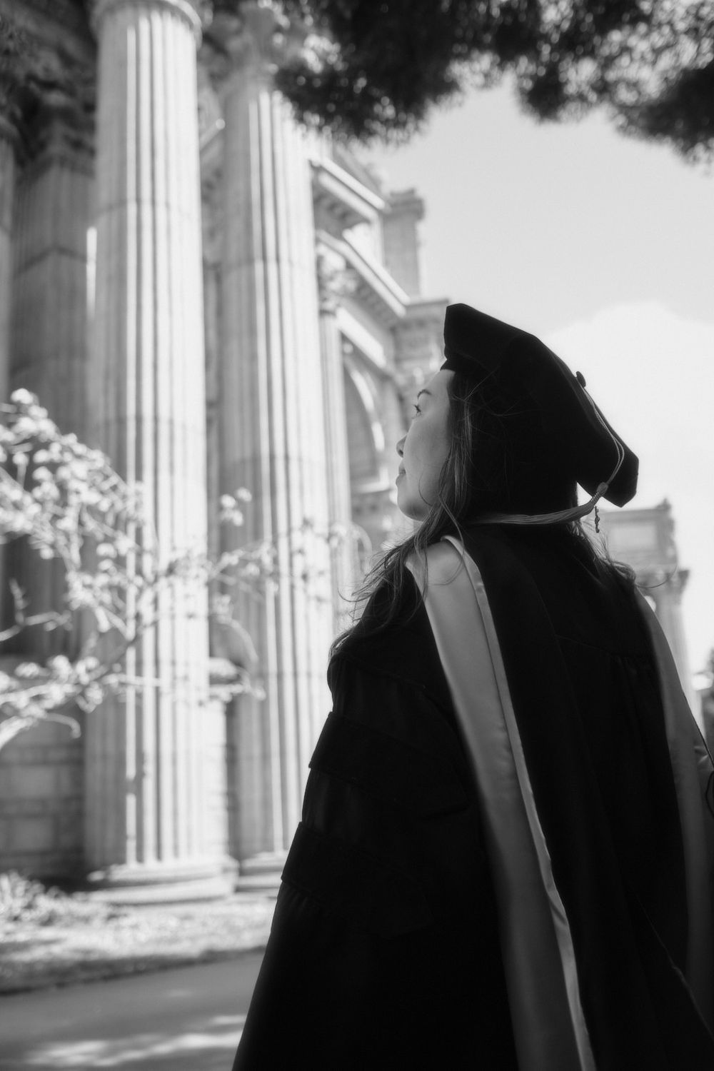 Black and white photo of graduate in cap and gown looking up at classical stone columns.