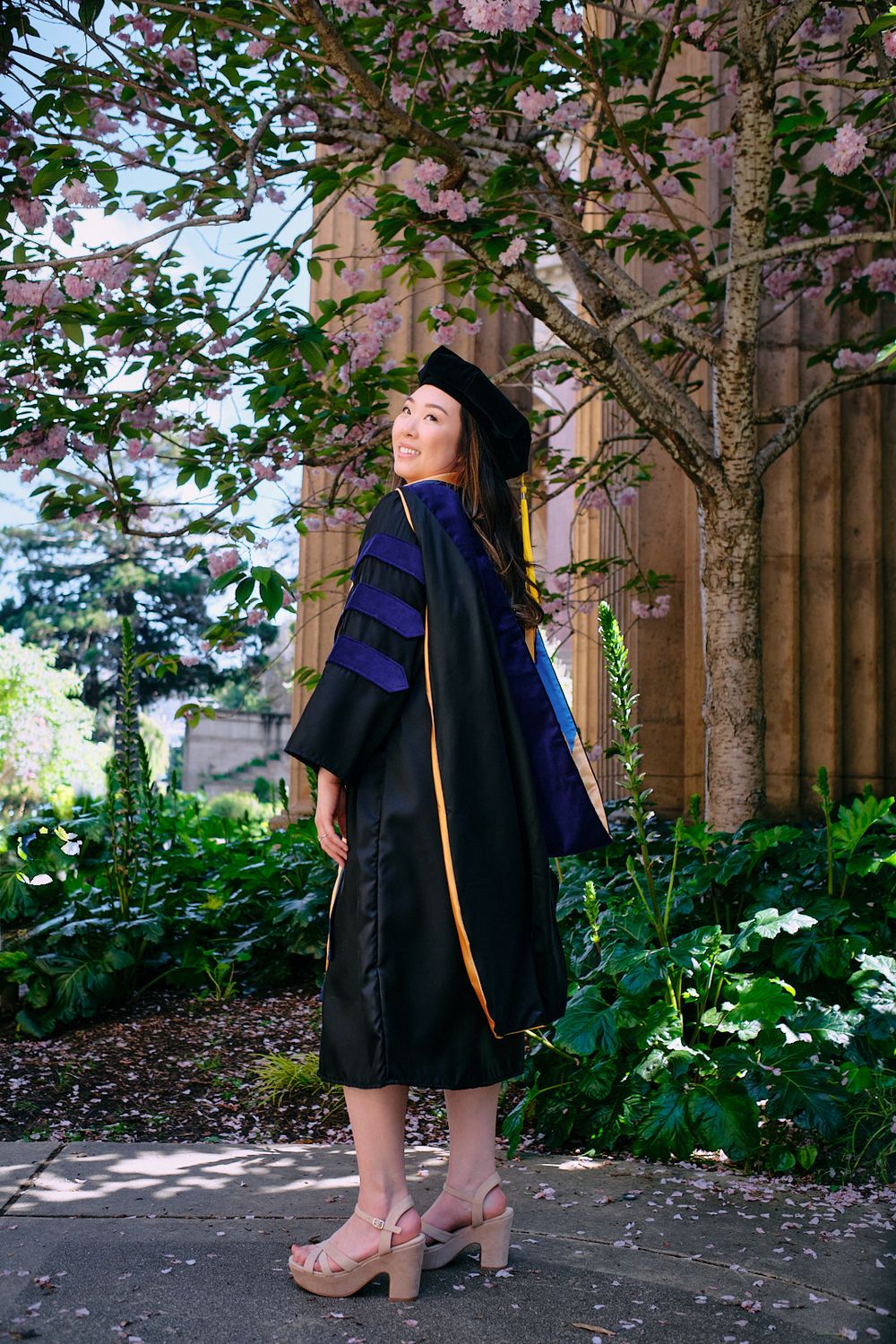 Graduate in doctoral cap and gown poses among pink cherry blossoms and lush green garden foliage.