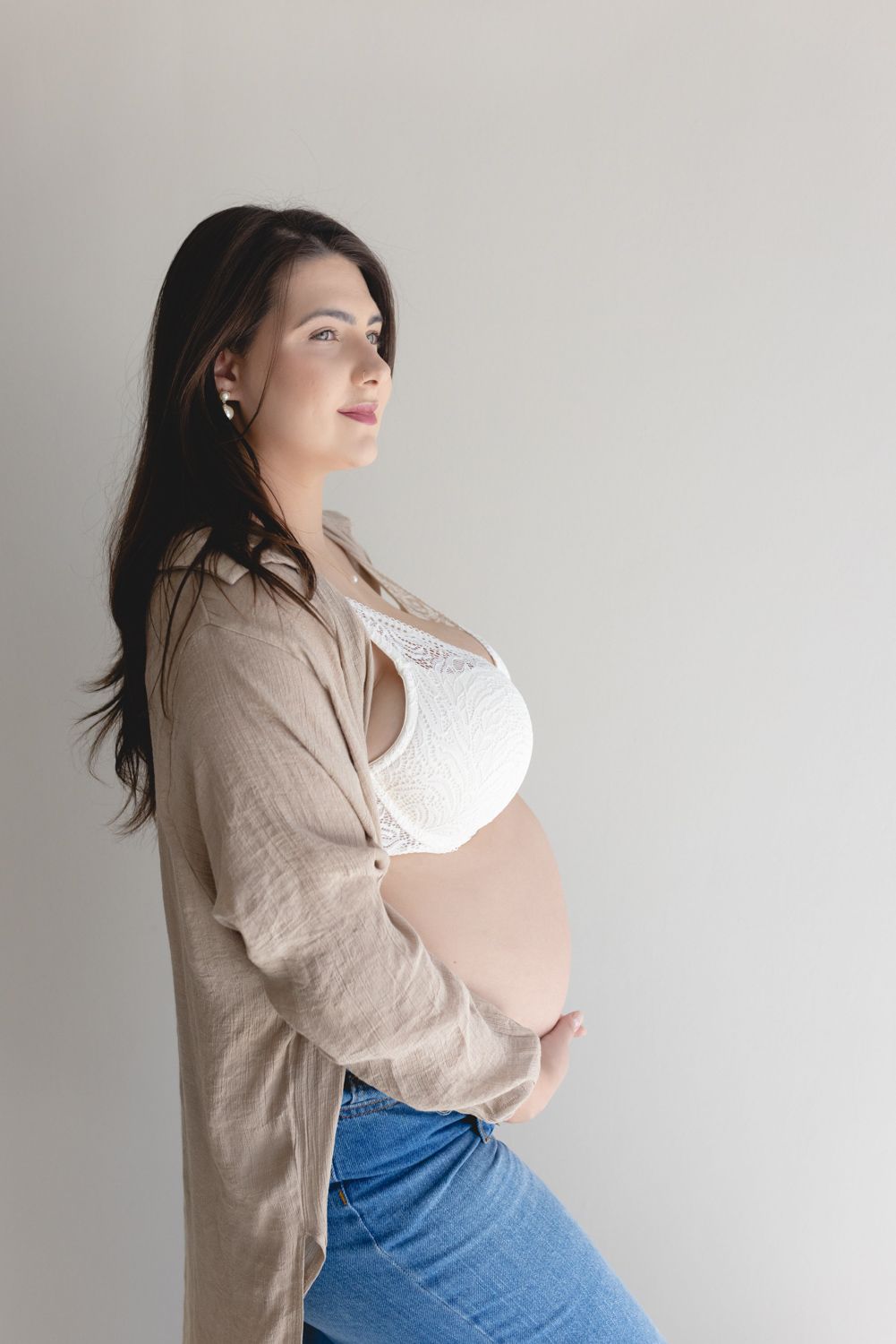 Expectant person in casual attire poses against white wall during pregnancy photoshoot.