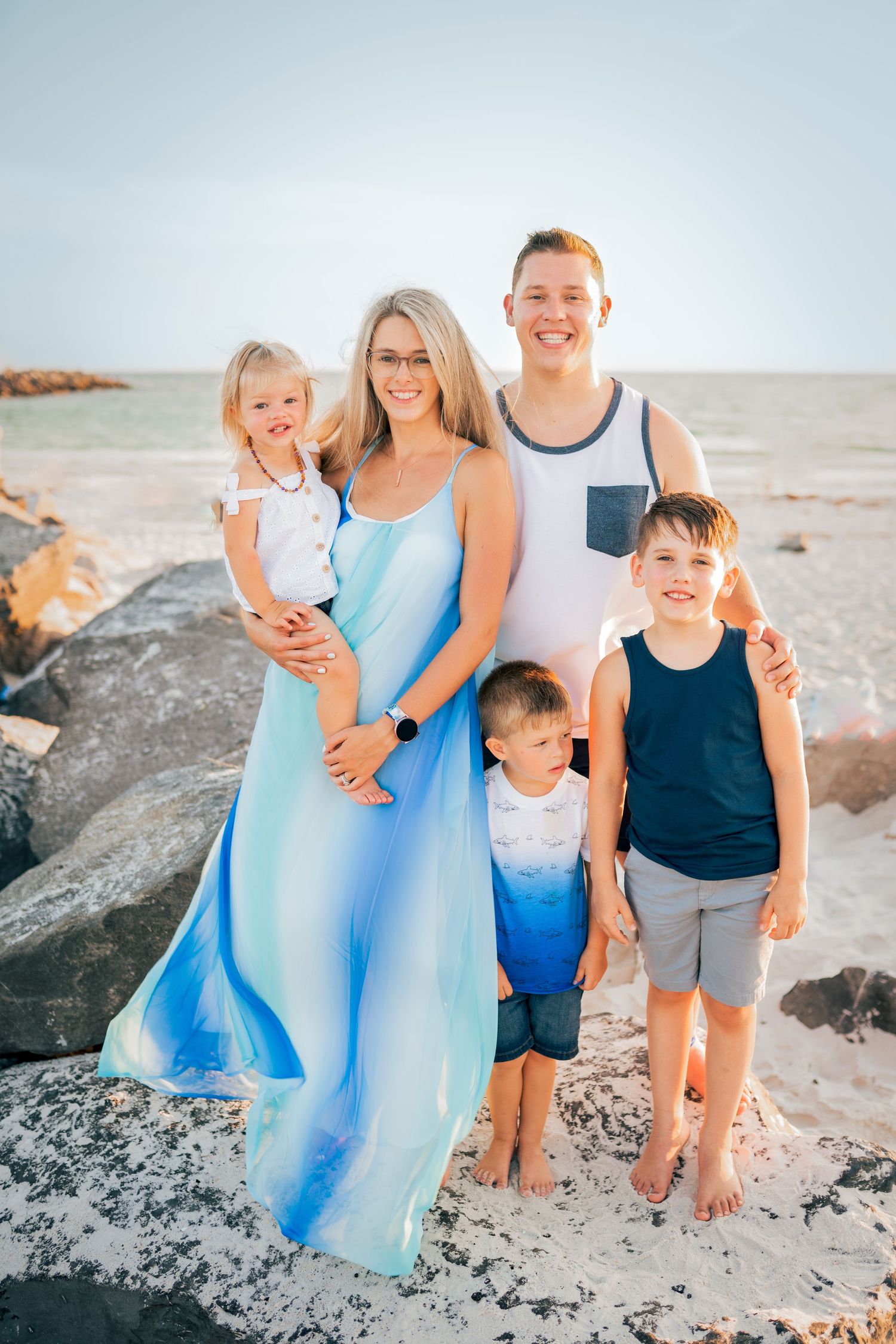 A family in coordinating blue outfits poses together on rocky beach during sunset.