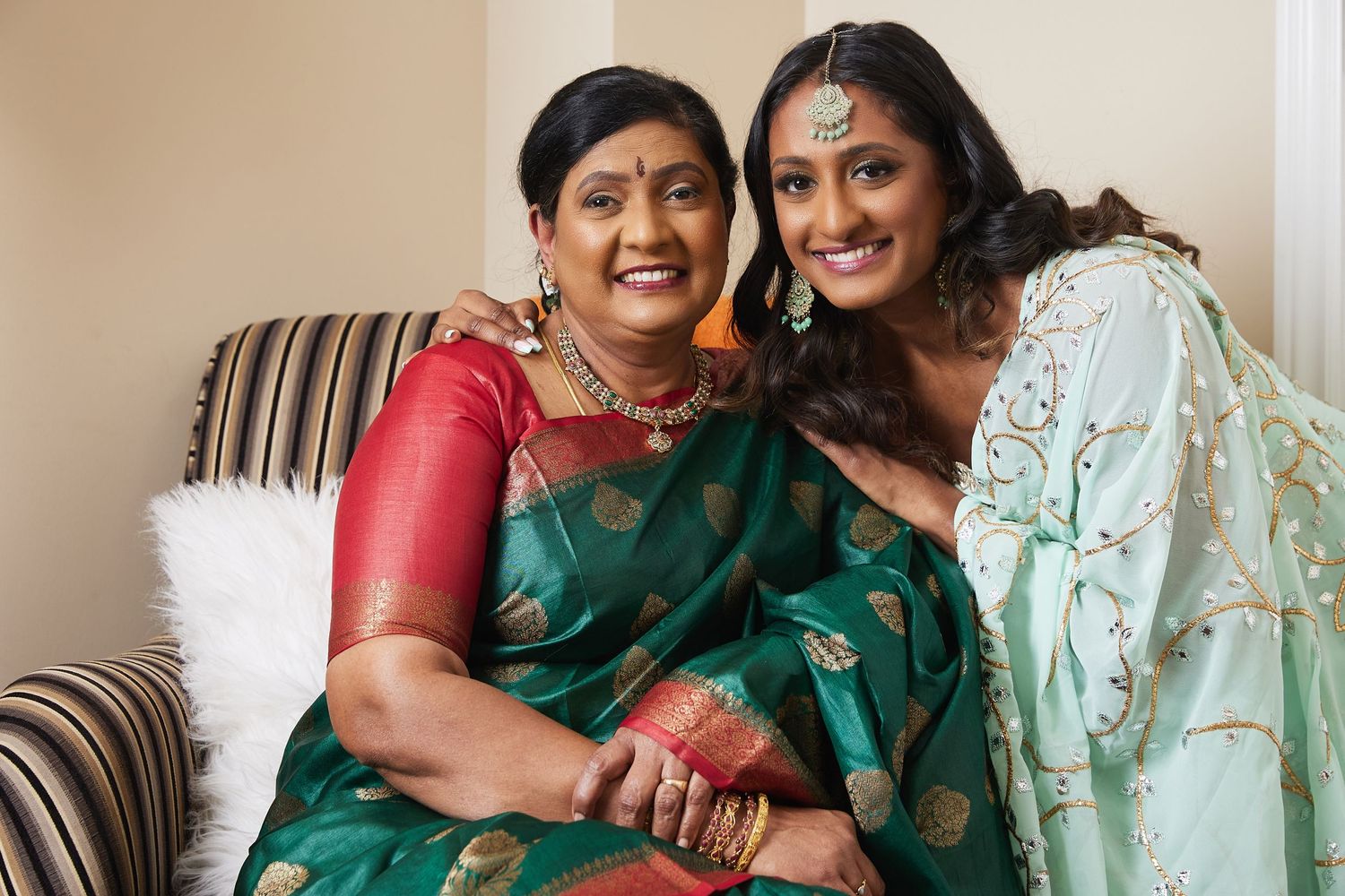Two women in traditional Indian saris sit close together on a couch sharing a warm moment.