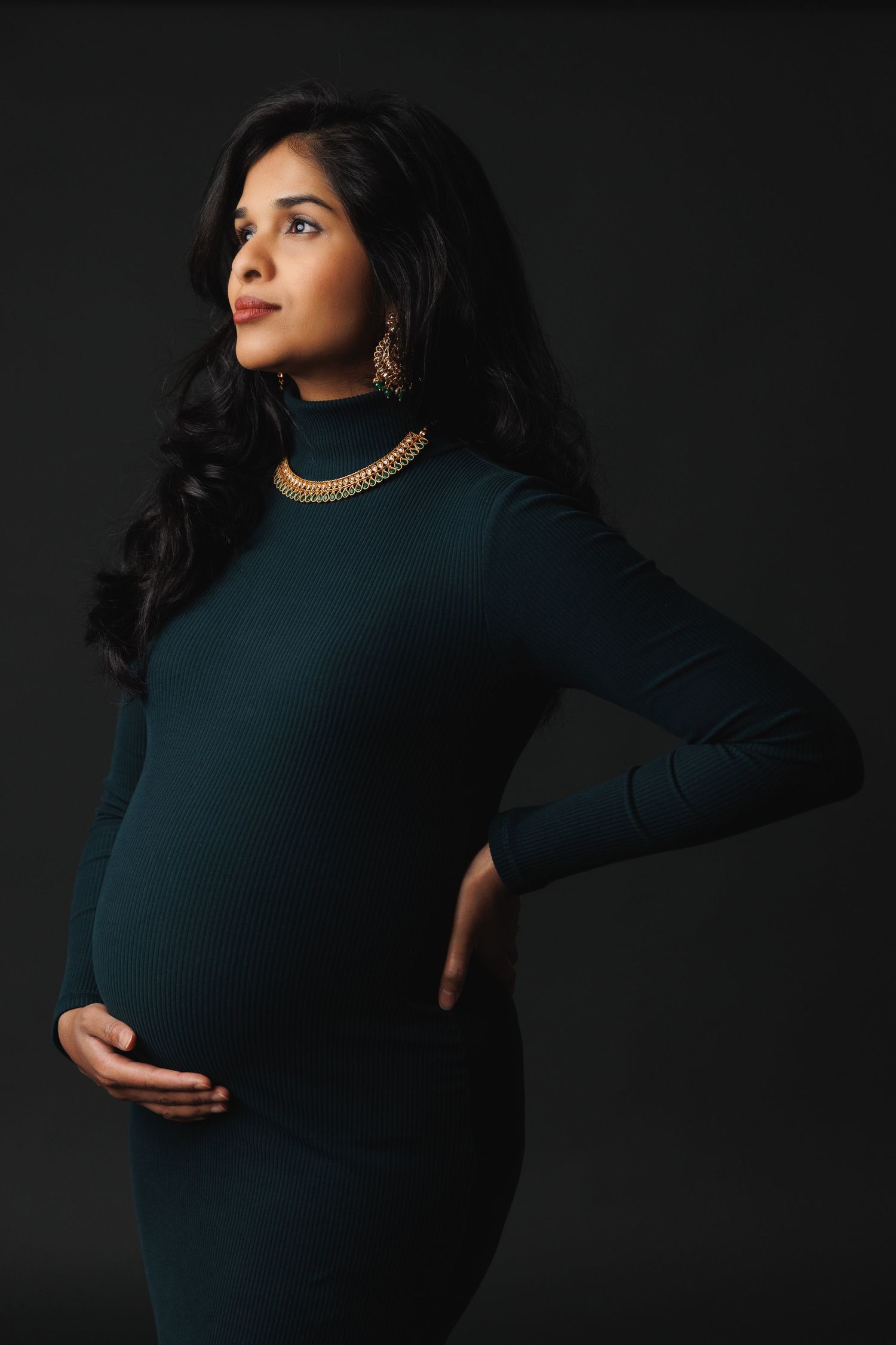 Elegant maternity portrait in teal dress against dark background with gold jewelry accents.