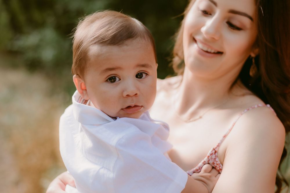 Mother in floral dress holding baby in white outfit against natural backdrop.