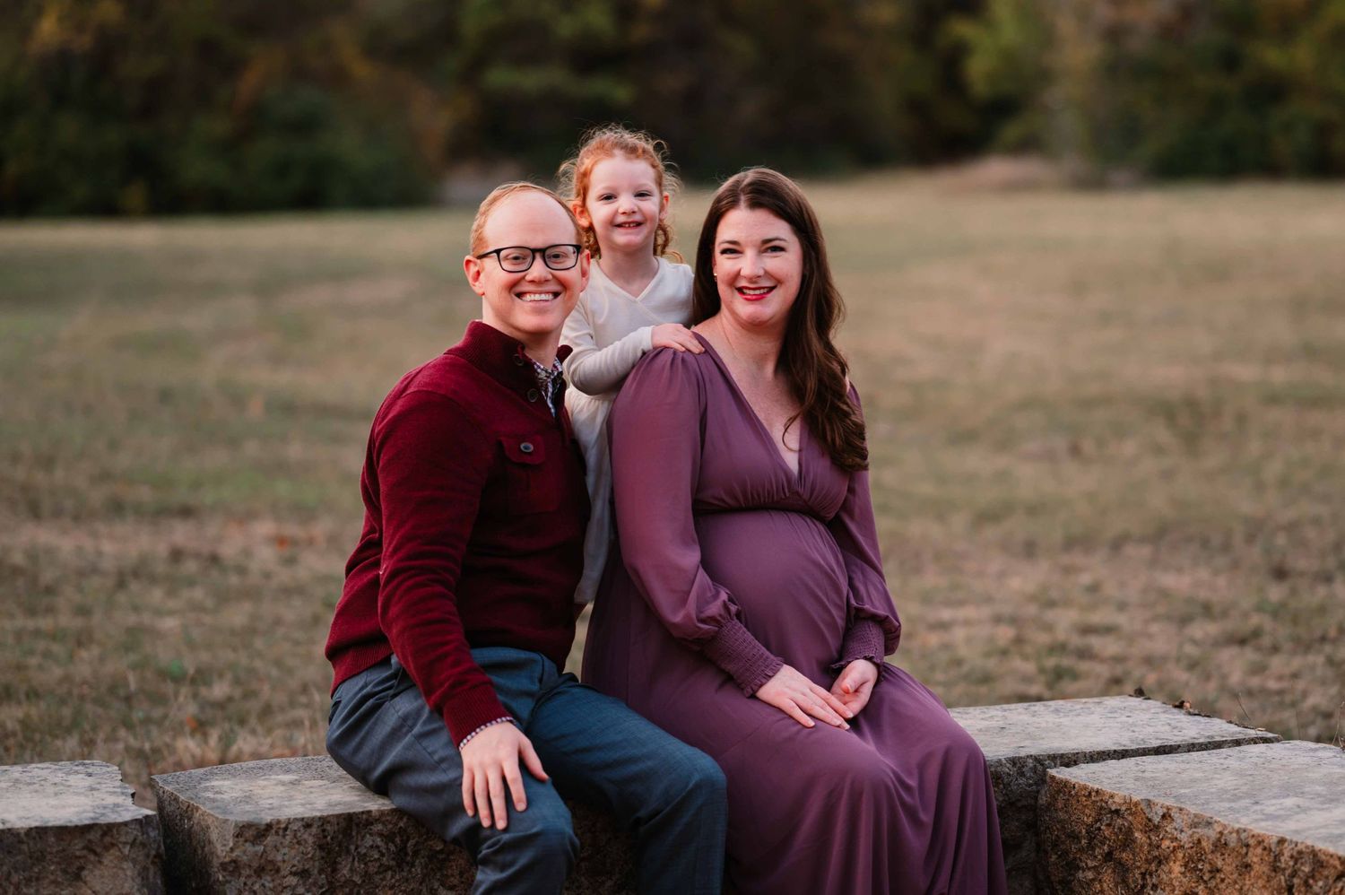 smiling family of pregnant mother, father, and preschool aged girl look at camera smiling during outdoor maternity photoshoot in dallas, tx