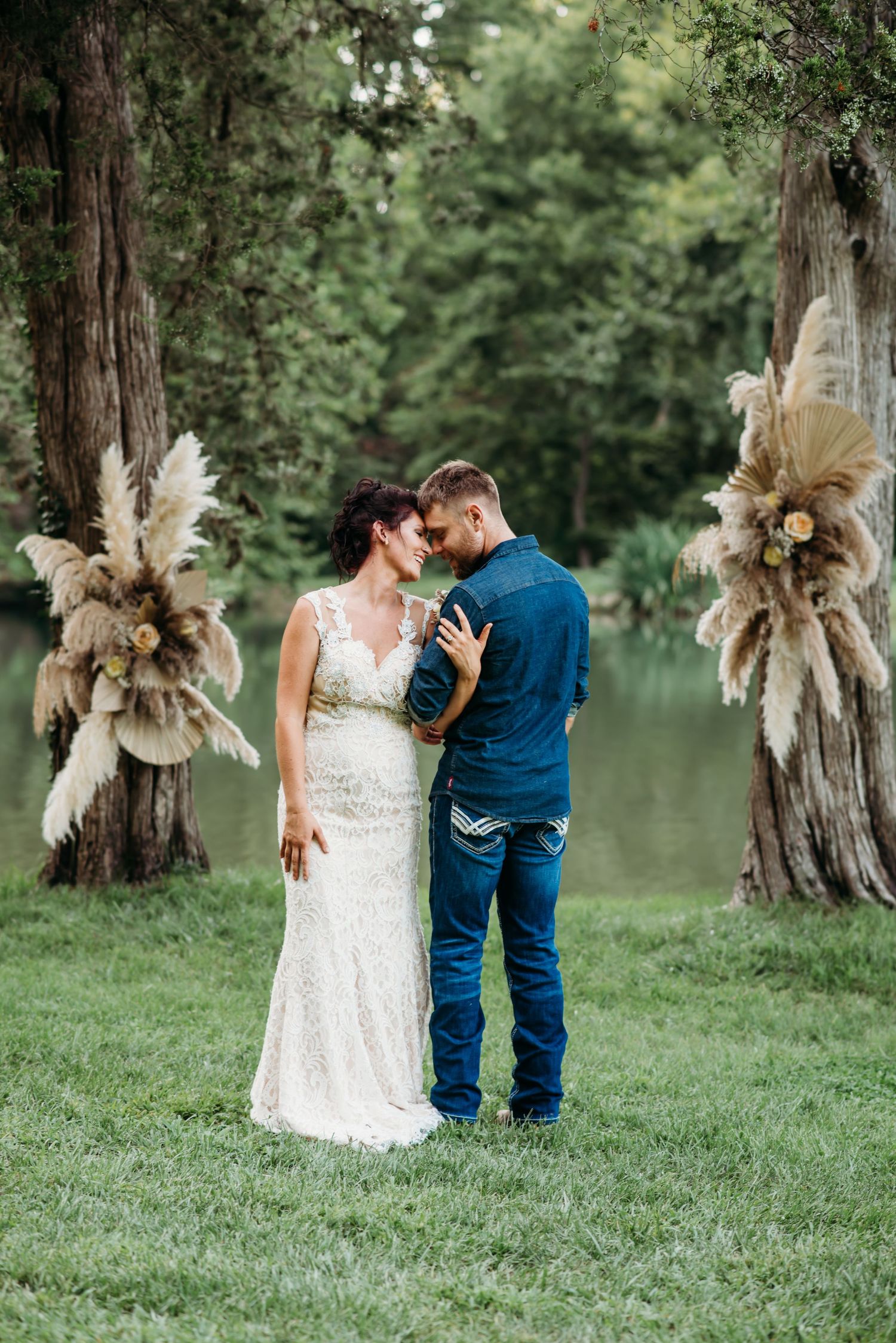 Couple in wedding attire embracing by lake with pampas grass decorations.