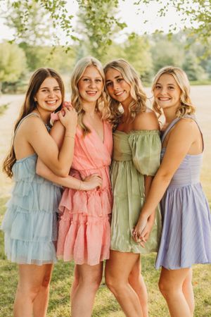 Four friends in pastel summer dresses pose together outdoors on a sunny day.