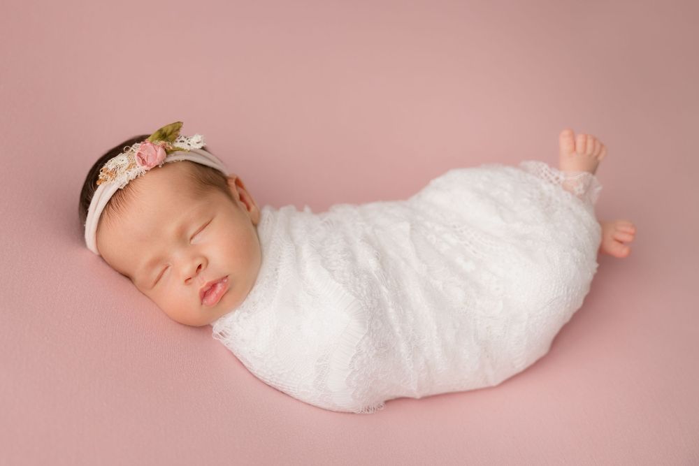 Sleeping newborn wrapped in white fabric wearing floral headband on pink backdrop.