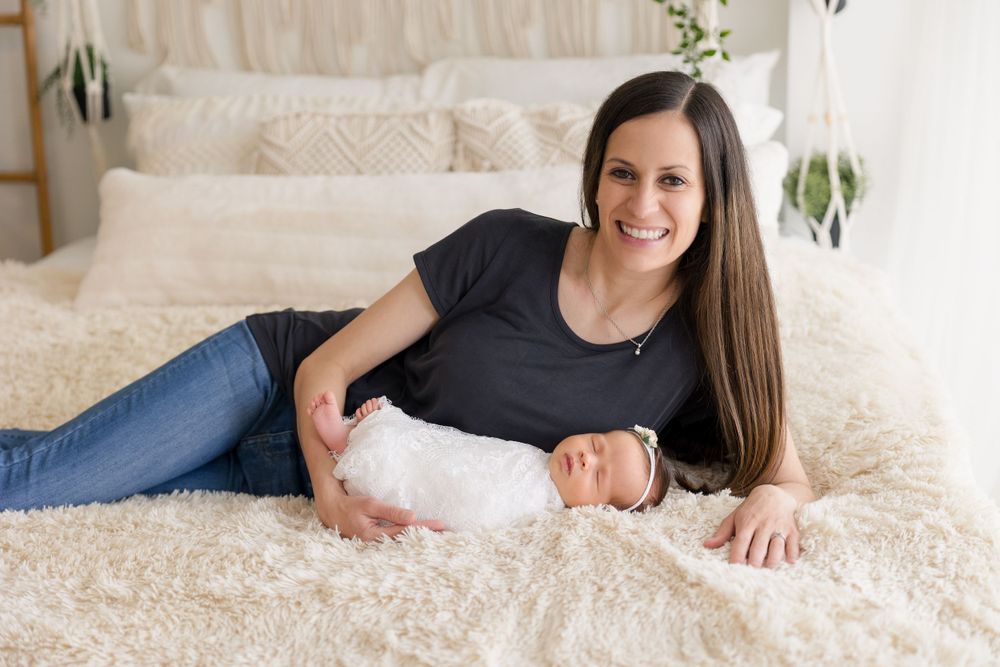 A parent and newborn relax together on a cozy white bed with decorative plants in background.