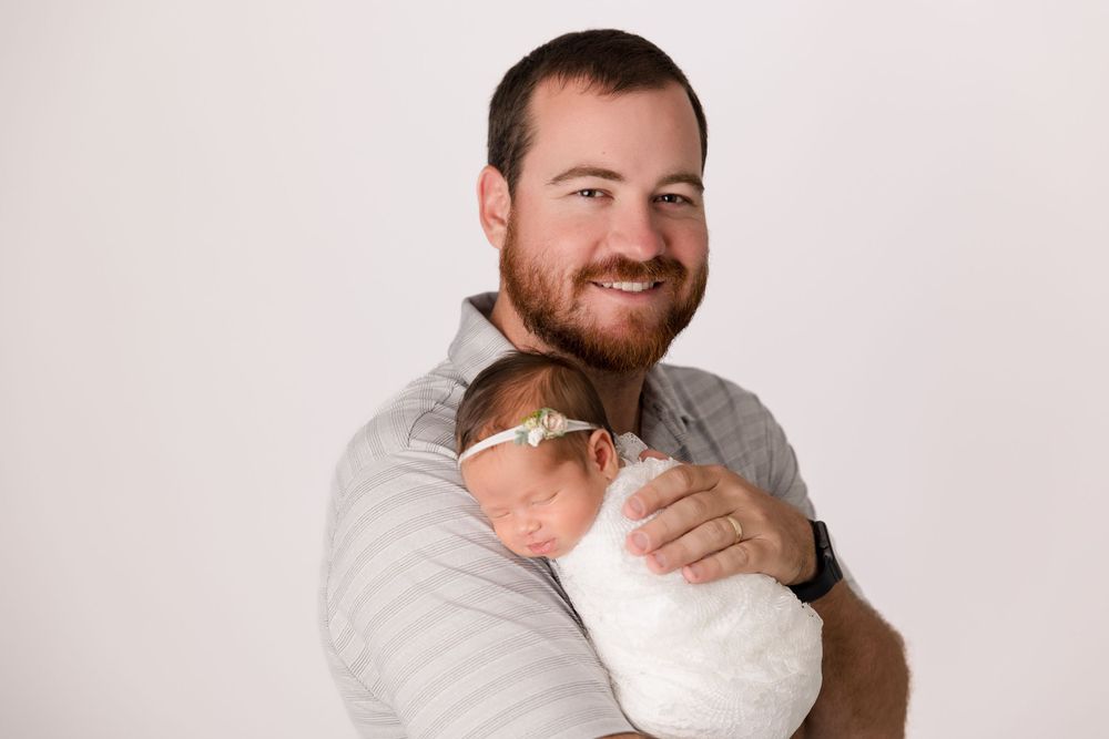 A parent gently cradles a sleeping newborn wrapped in white against a neutral backdrop.