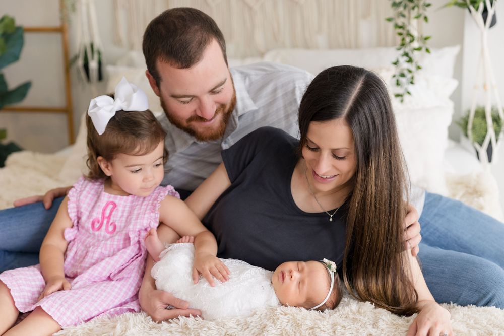 Family of four relaxes together on white bedding with greenery in background.