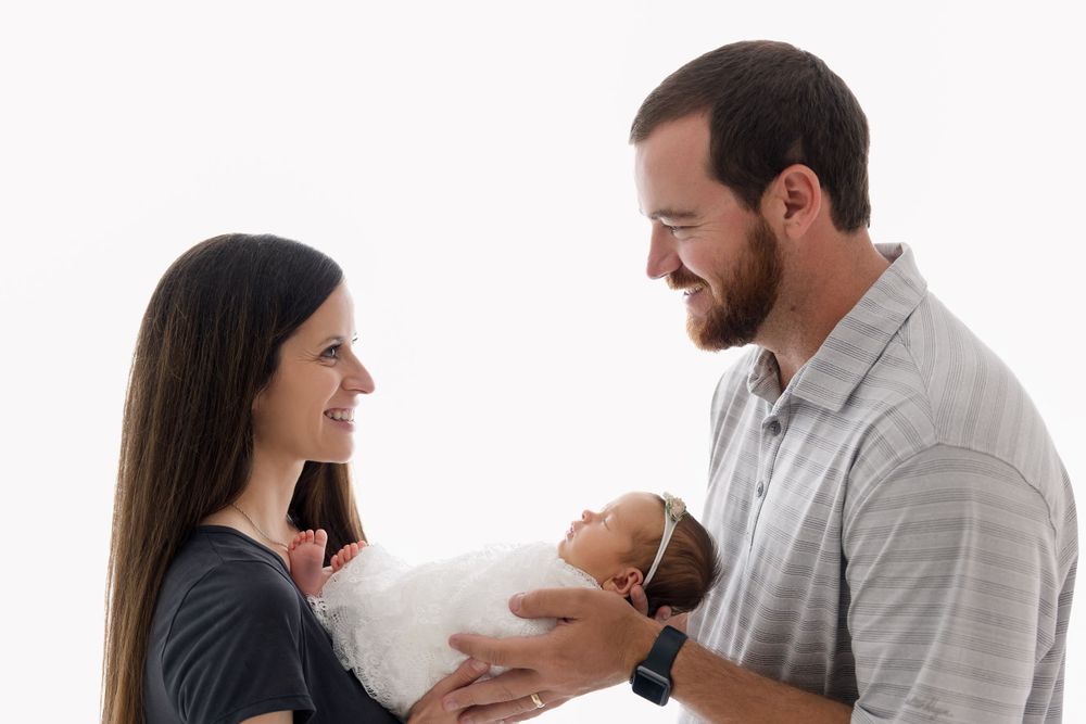 Parents sharing a tender moment while holding their newborn baby against white backdrop.