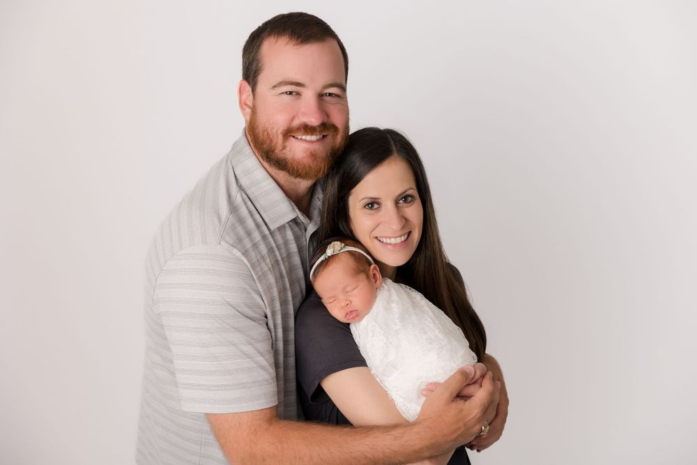 A loving couple embraces while holding their newborn baby against a light background.