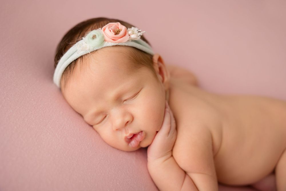 Peaceful newborn with pink floral headband resting on soft pink backdrop.