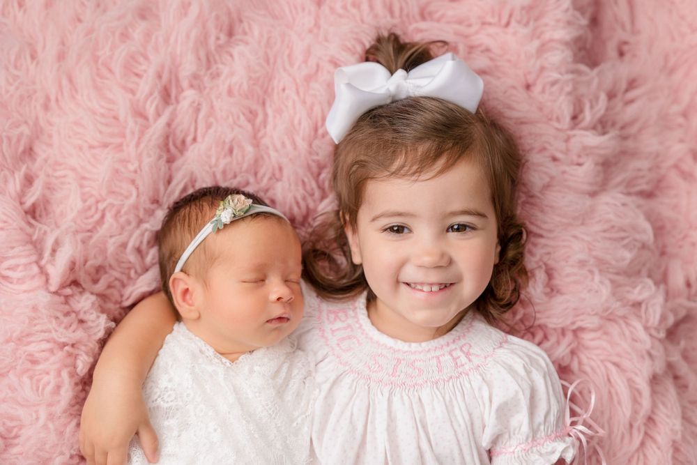 Two young children in white outfits lying on pink fluffy backdrop.