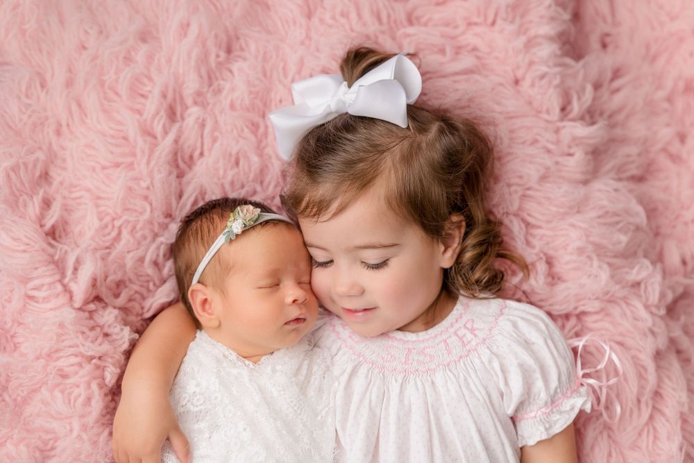Two siblings in white dresses rest together on pink fluffy backdrop.