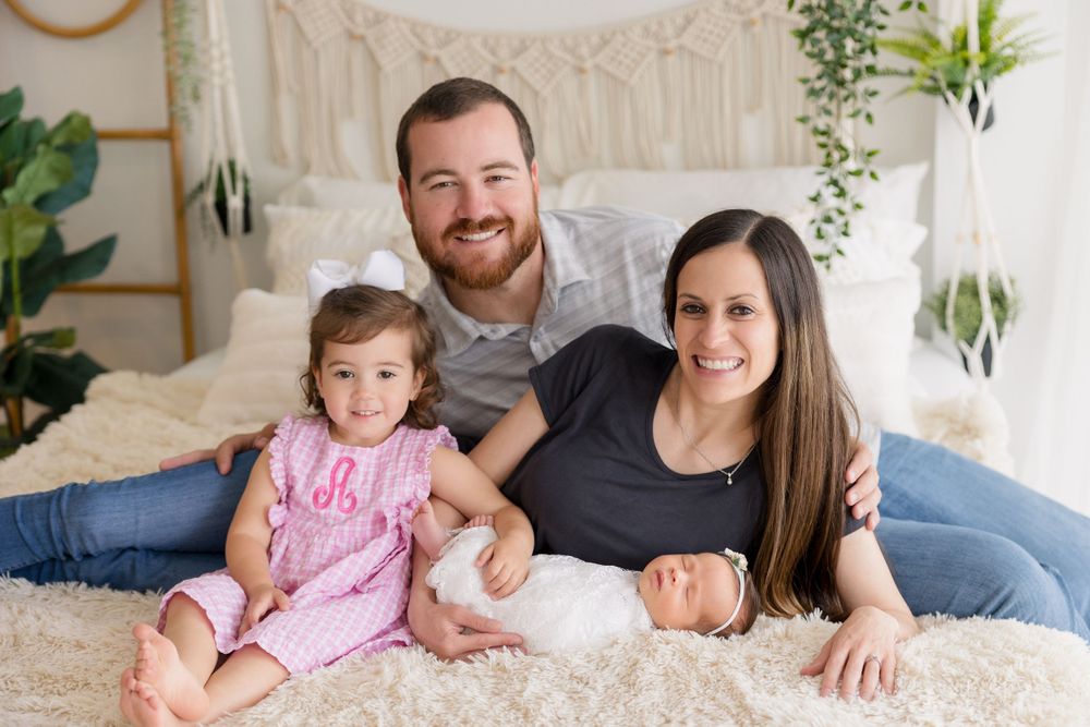 Happy family of four poses together on white bed with macrame wall hanging in background.