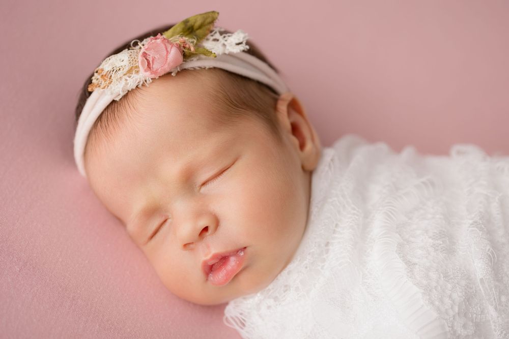 Peaceful sleeping newborn wearing floral headband on pink background.