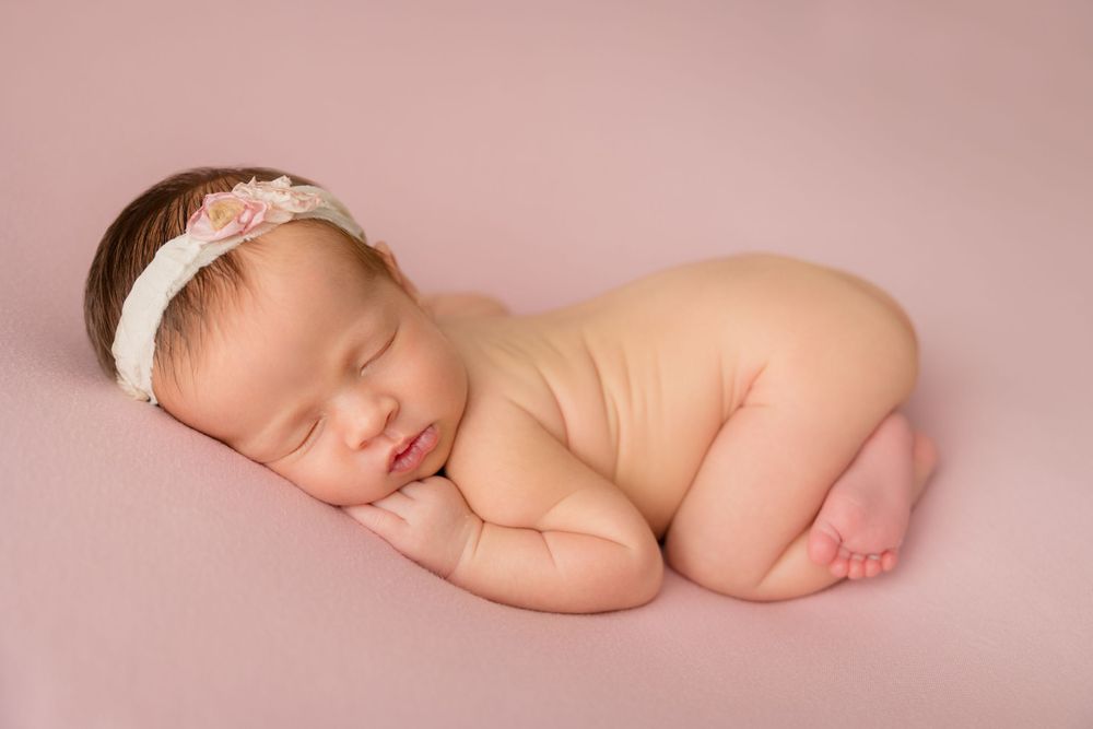Newborn baby sleeping in fetal position with floral headband on pink background.