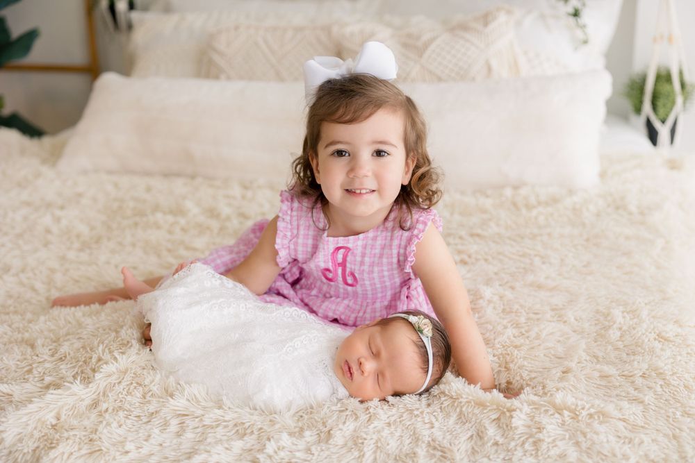 Young child in pink dress sits with baby sibling on white bedding.