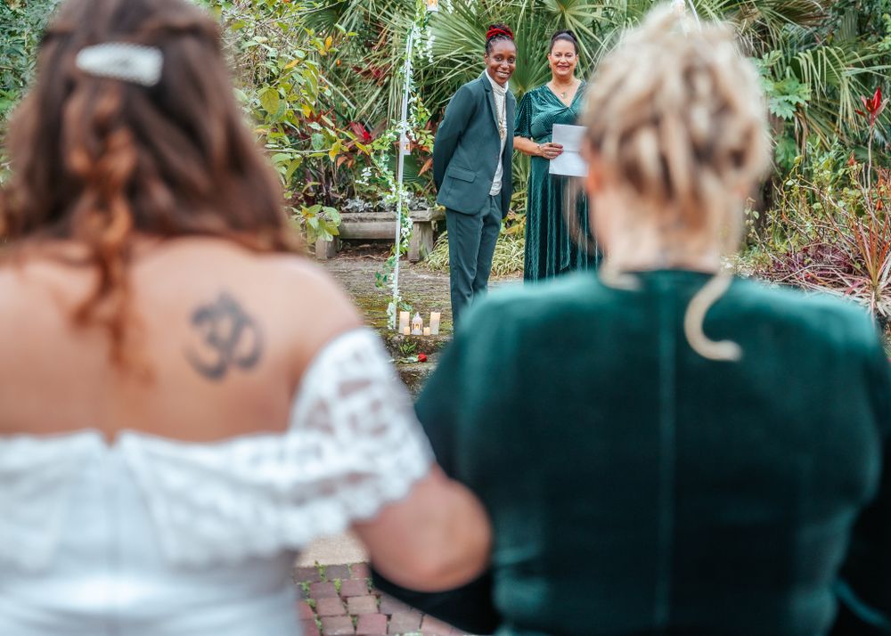 Wedding ceremony photographed from behind guests with couple in green attire visible in background.