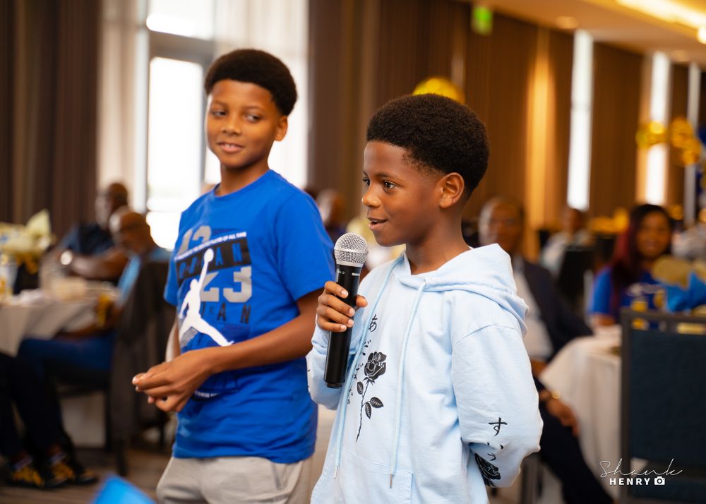 Two young boys performing at a community event, one holding a microphone while wearing a light colored shirt.