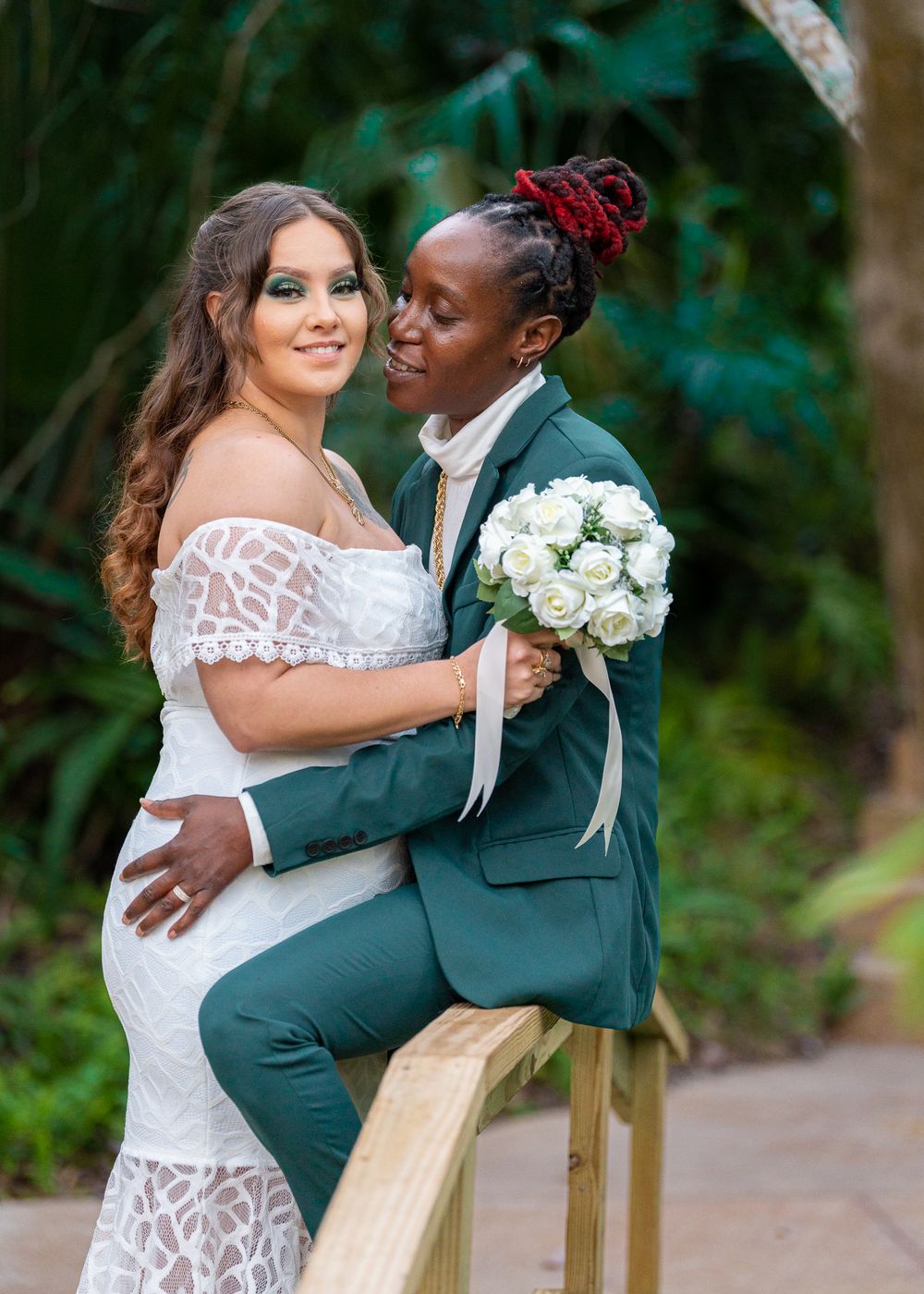 Romantic wedding portrait featuring a couple in white dress and dark green suit holding white roses in an outdoor garden setting.