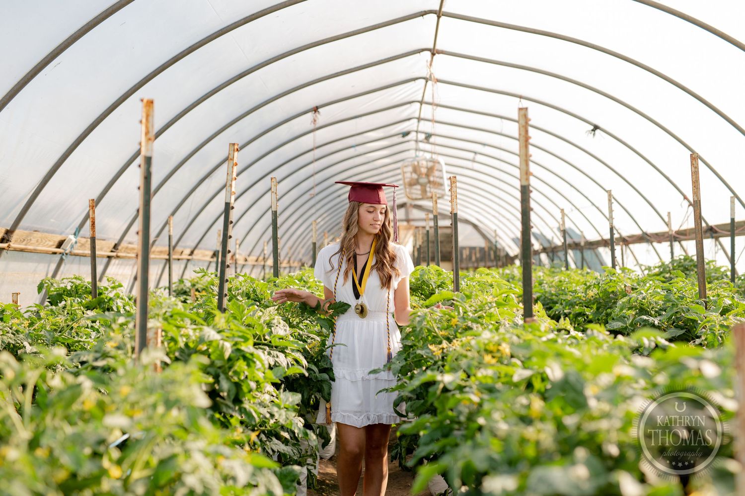 Graduate in white dress stands amid flourishing greenhouse plants under curved white ceiling.