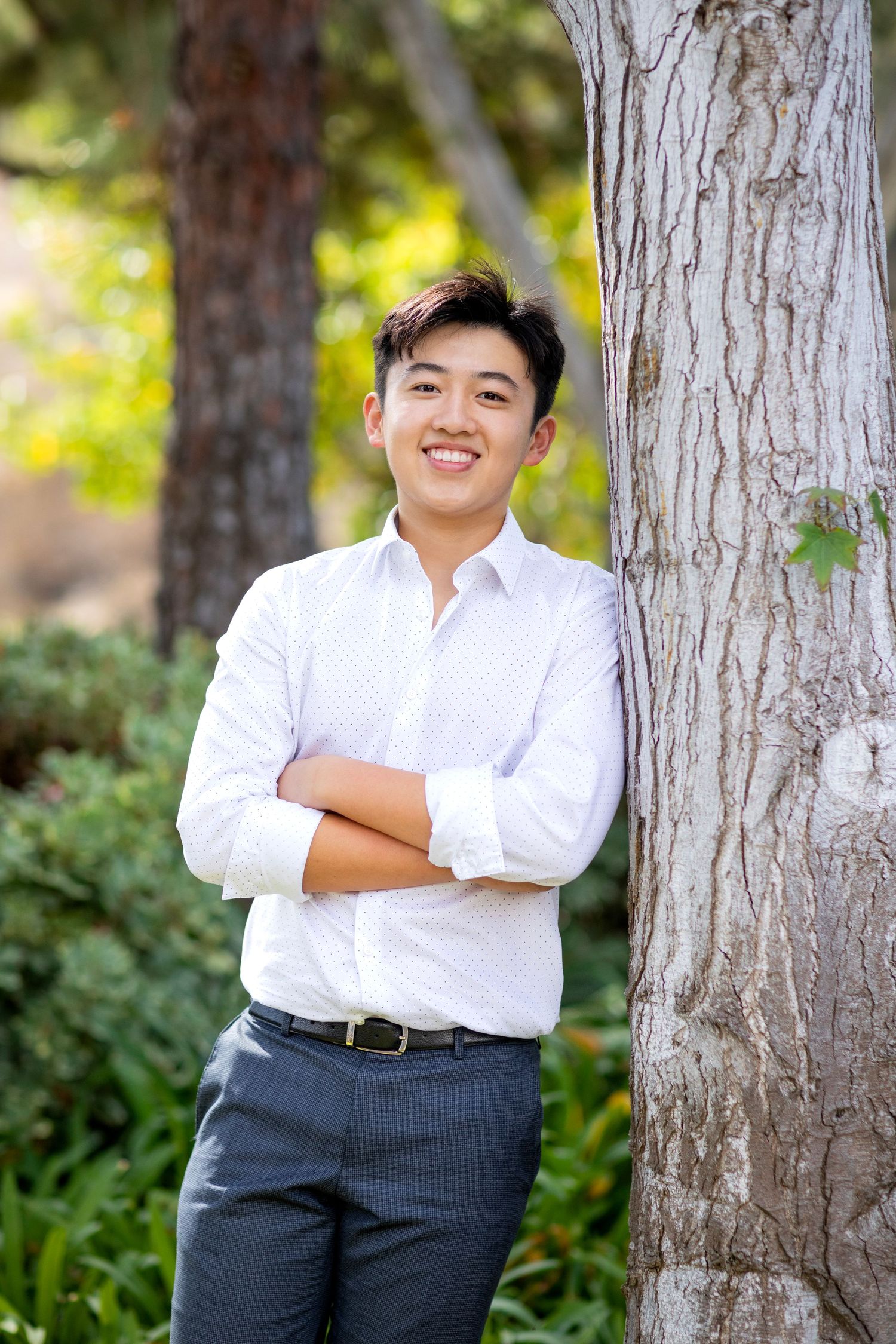 Young person in white dress shirt and dark pants poses confidently against tree trunk in outdoor nature setting.