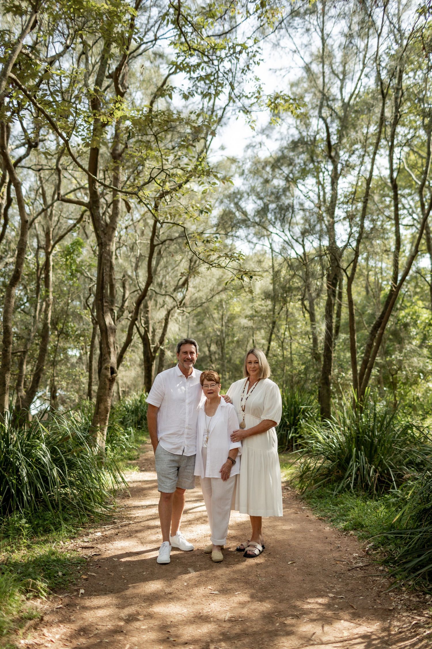 Three people in light colored clothing walk together on a tree-lined dirt path.