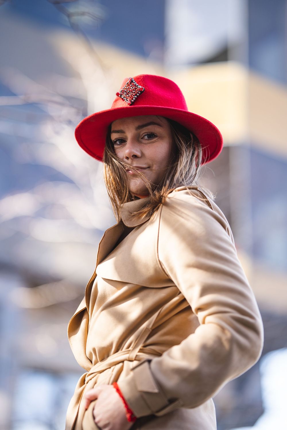 Stylish woman in beige coat and red hat with embellishment poses against blurred background.