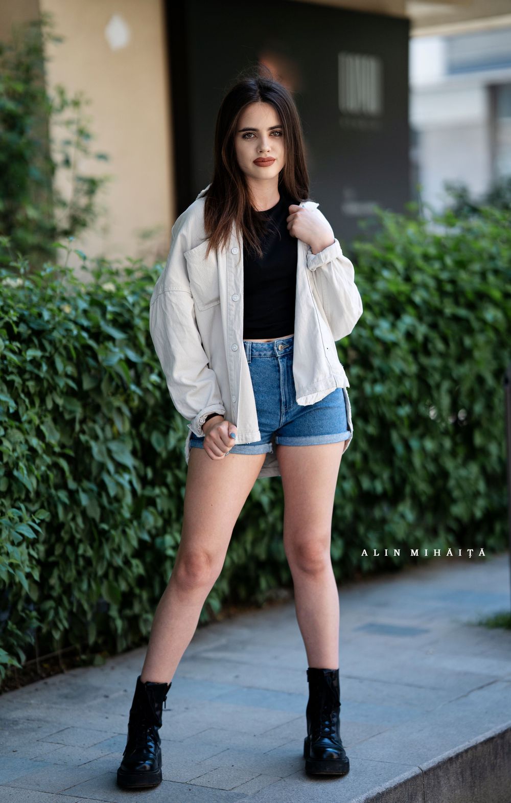 Casual summer outfit with white blouse, denim shorts and black combat boots against green foliage.