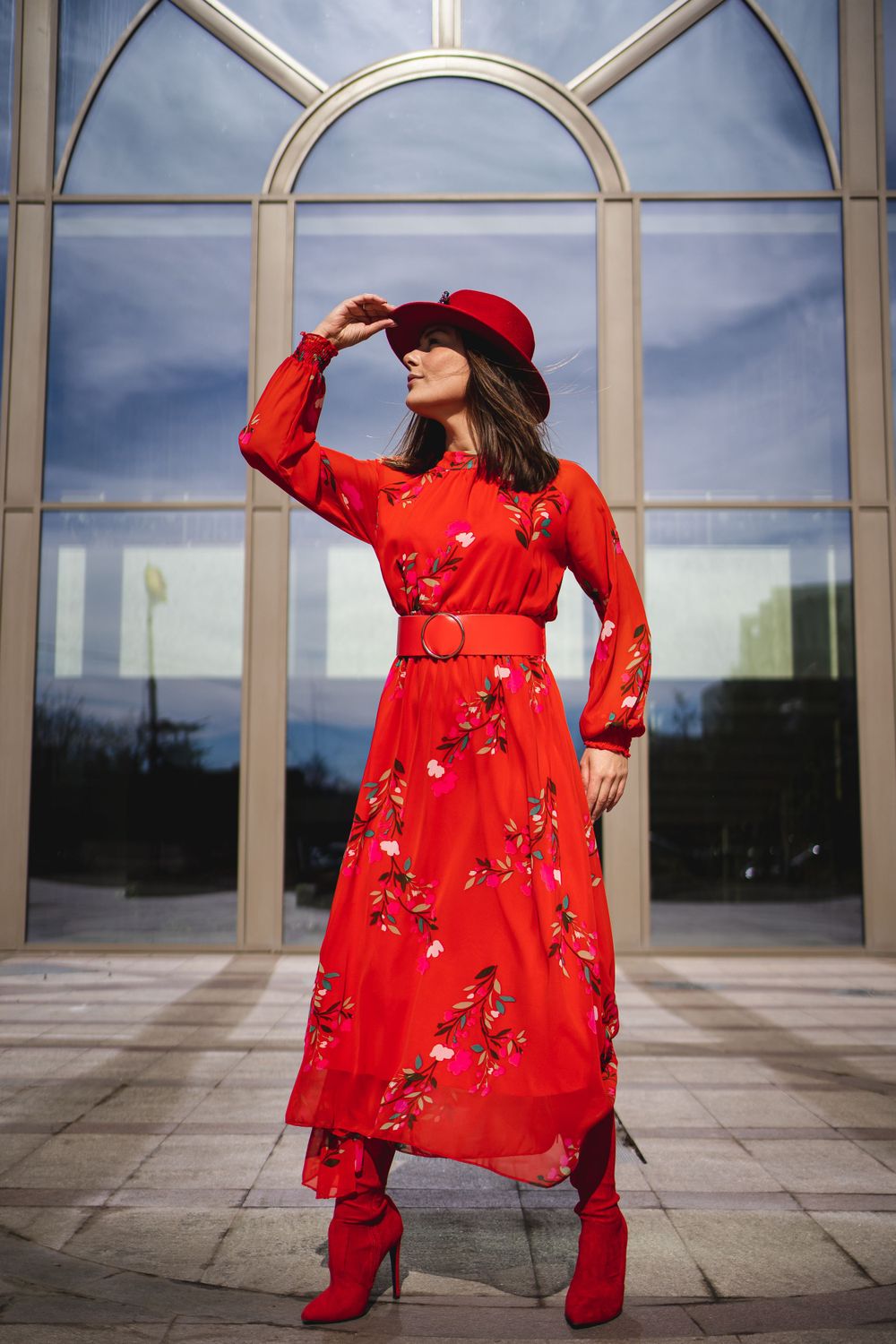 Woman in flowing red floral dress and hat poses dramatically under architectural archway.