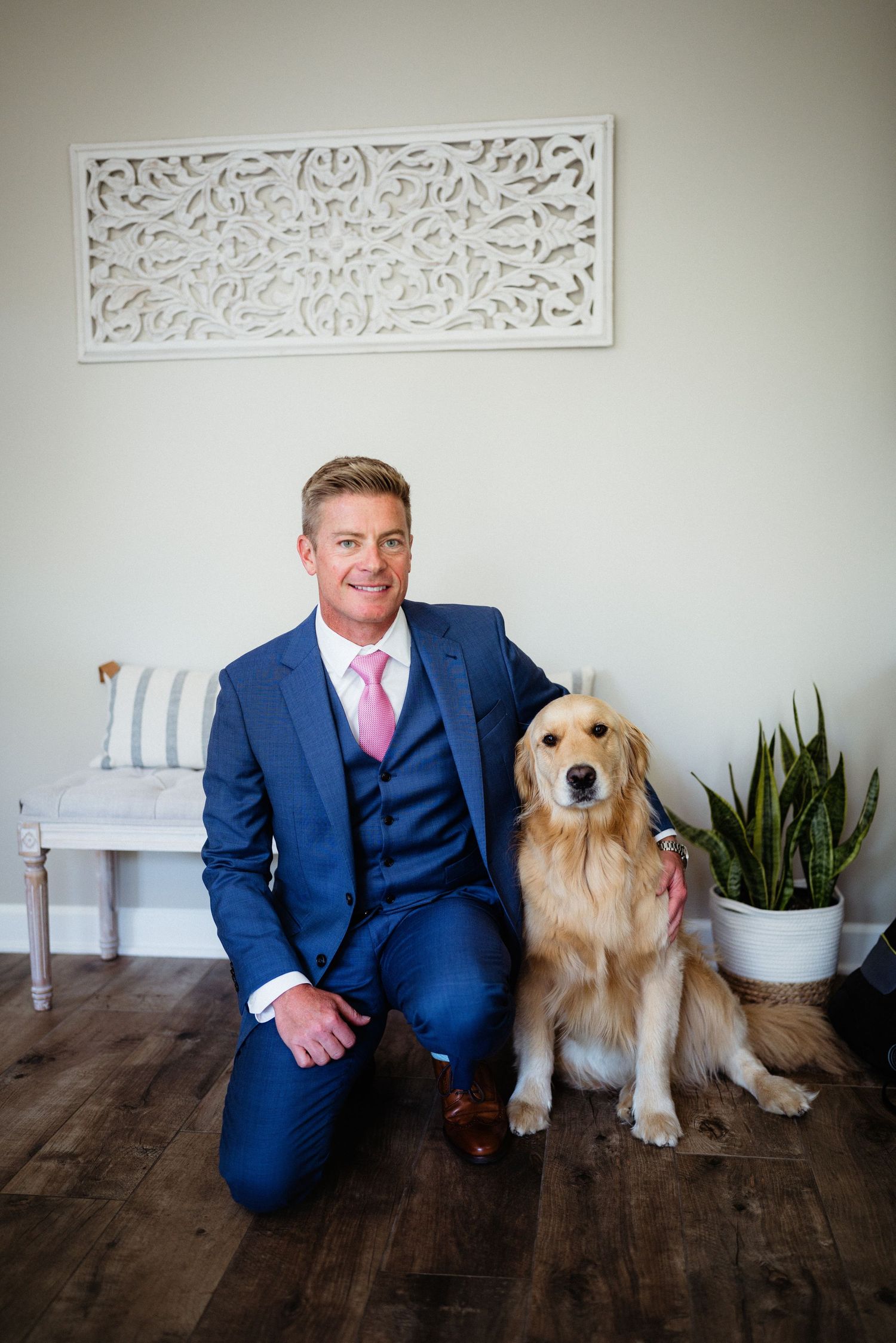 A Golden Retriever sits next to someone in a navy suit against a white wall with decorative artwork.