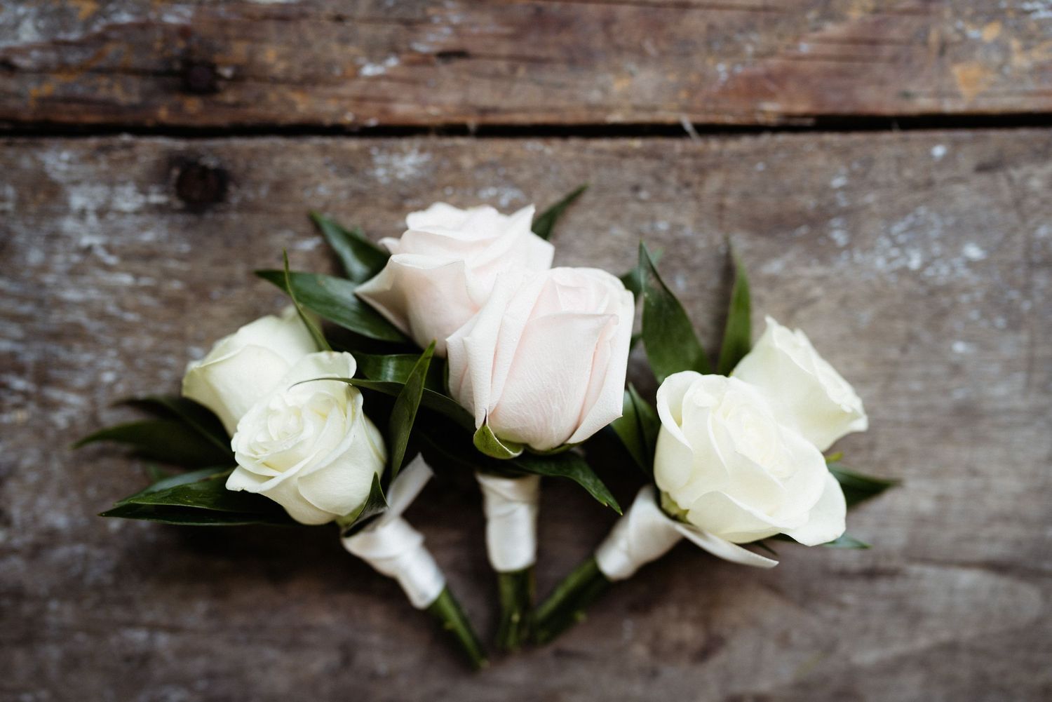 Three delicate white and pink rose boutonnieres arranged on rustic wooden surface.