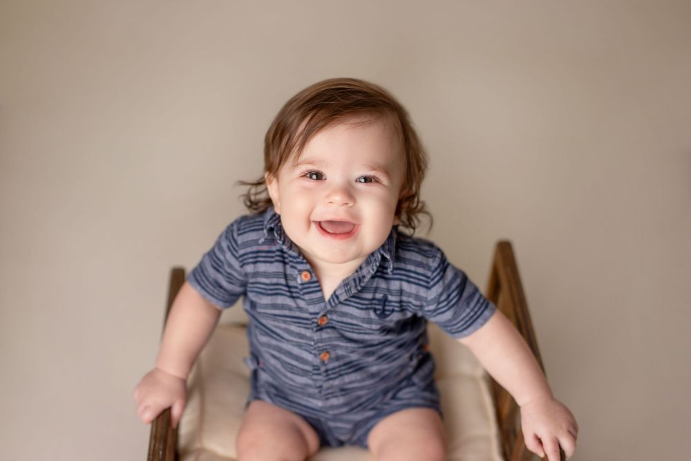 Baby in striped polo onesie sitting in wooden chair.