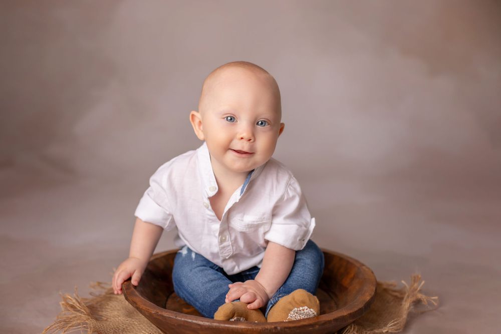 Baby in white shirt sits in wooden bowl with rustic backdrop.