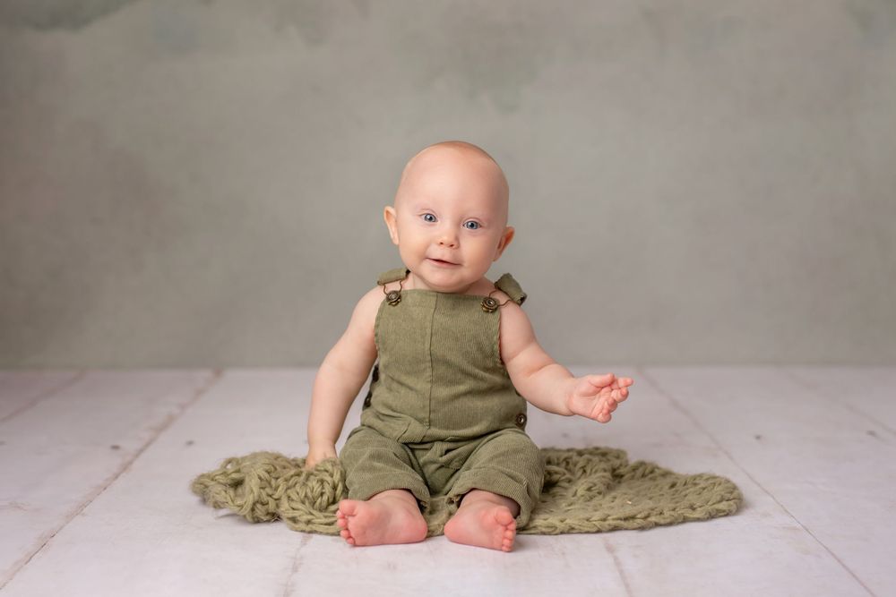 Baby wearing olive green romper sitting on textured rug.