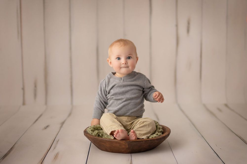 Baby in grey shirt sits in wooden bowl on white surface.