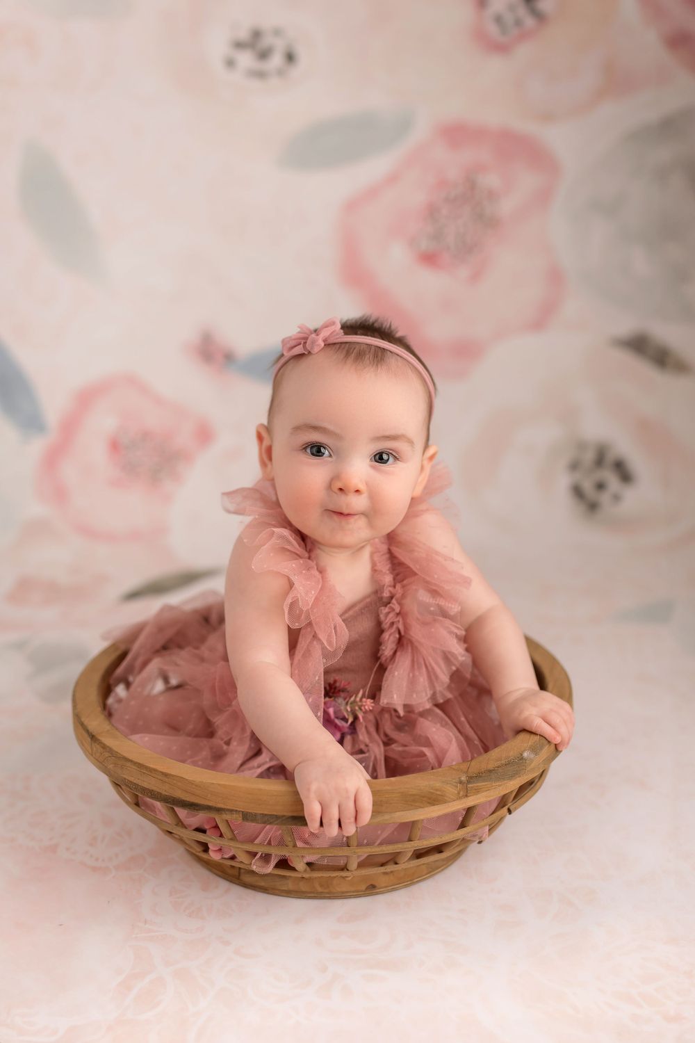 Baby in pink tulle dress sitting in wooden basket against soft floral background.