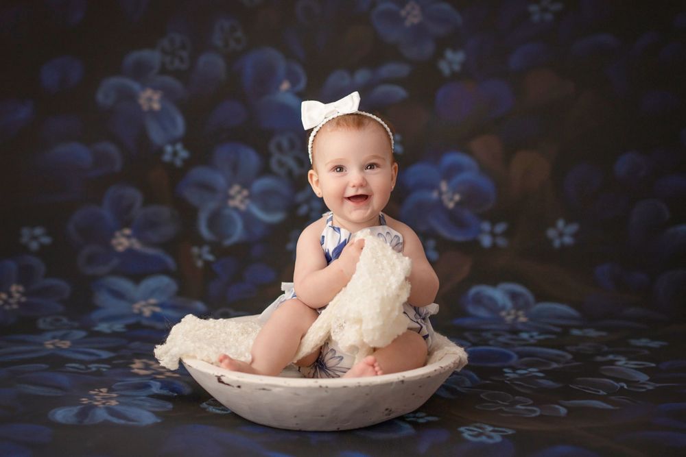 Baby in white dress sits in vintage bowl against blue floral backdrop.