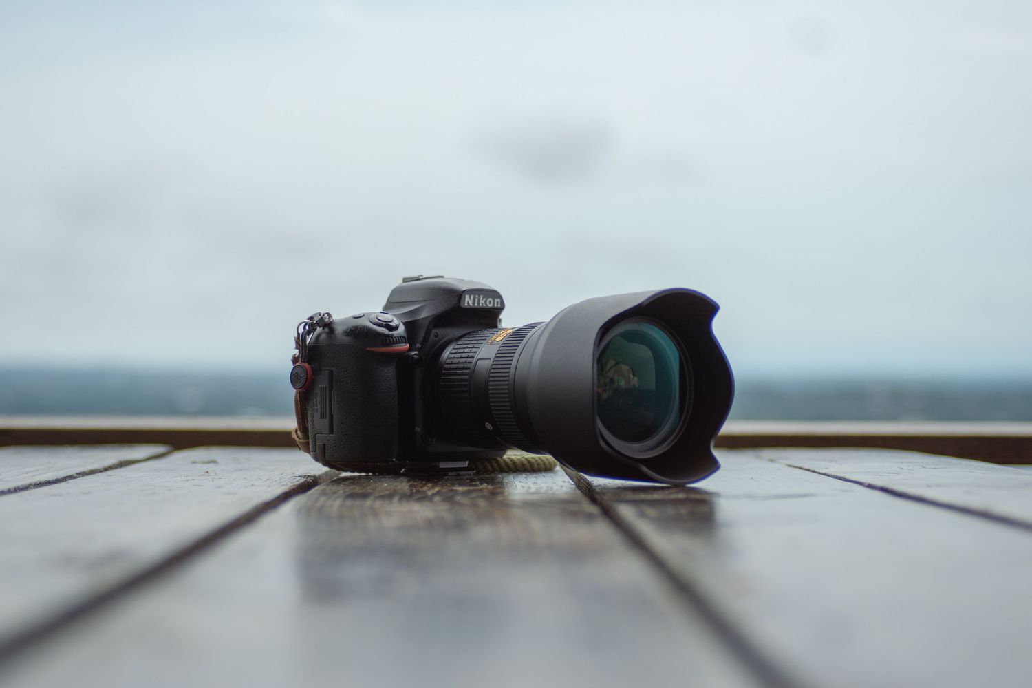 A black DSLR camera with telephoto lens sitting on wooden planks against an overcast sky.